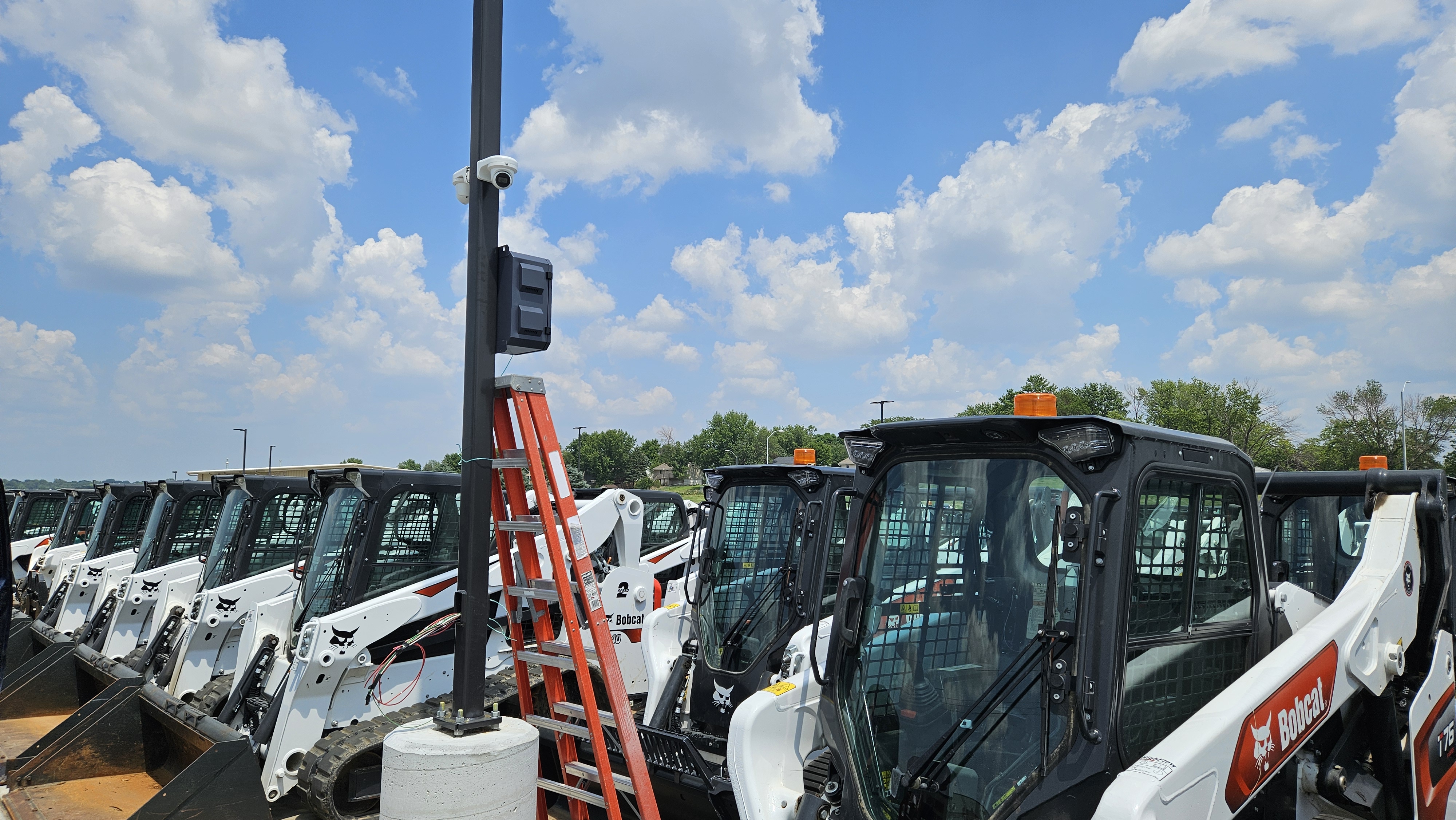 Security camera pole installation among rows of Bobcat skid steers at equipment dealership